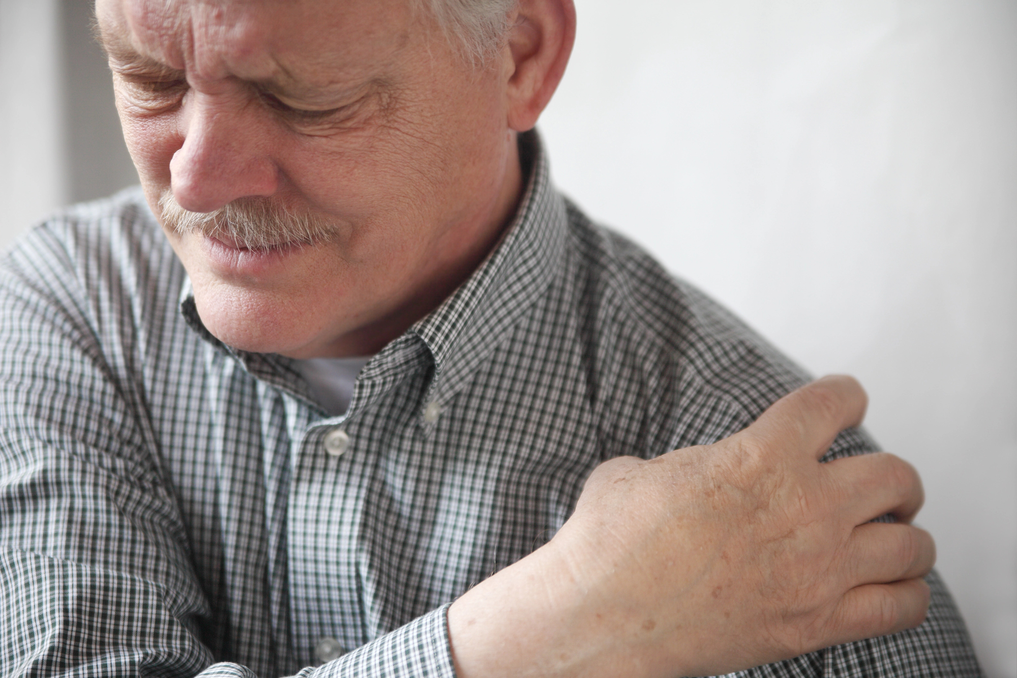 A man with a mustache is wearing a checkered shirt and clutching his left shoulder with a pained expression, suggesting discomfort or pain in that area.