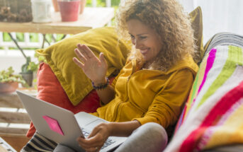 A woman with curly hair, wearing a yellow hoodie, is sitting on a couch with colorful pillows and a blanket. She is smiling and waving at a laptop screen. A wooden table with plants is in the background.