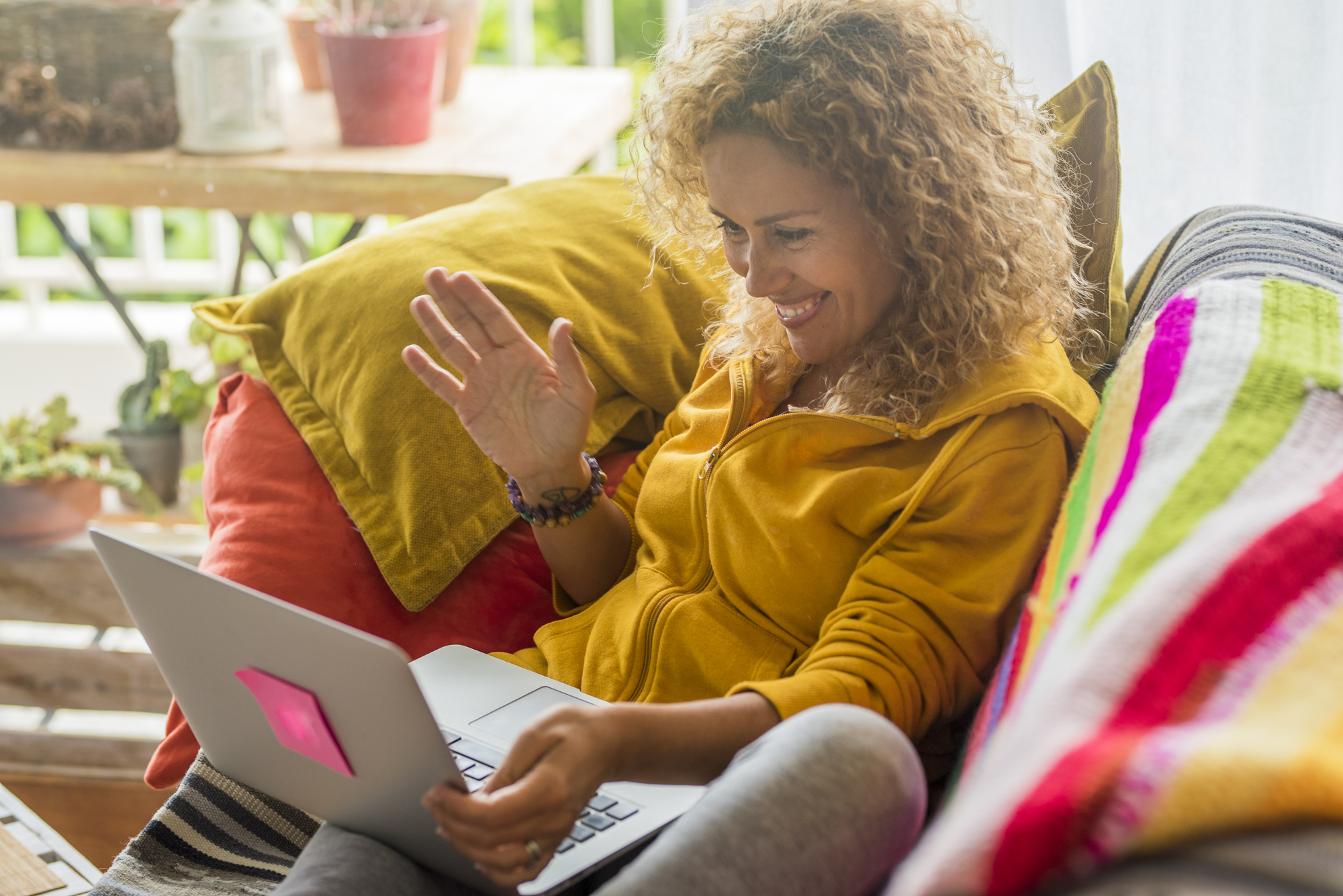 A woman with curly hair, wearing a yellow hoodie, is sitting on a couch with colorful pillows and a blanket. She is smiling and waving at a laptop screen. A wooden table with plants is in the background.