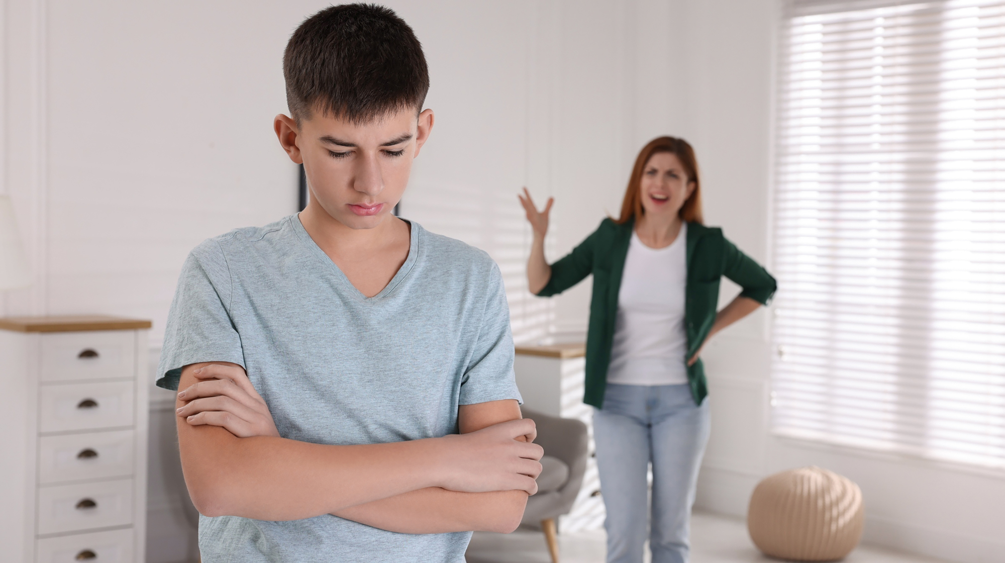 A teenage boy stands with arms crossed, looking down, while a woman in a green jacket gestures animatedly in the background. They are in a brightly lit room with white walls and furniture. The atmosphere appears tense.