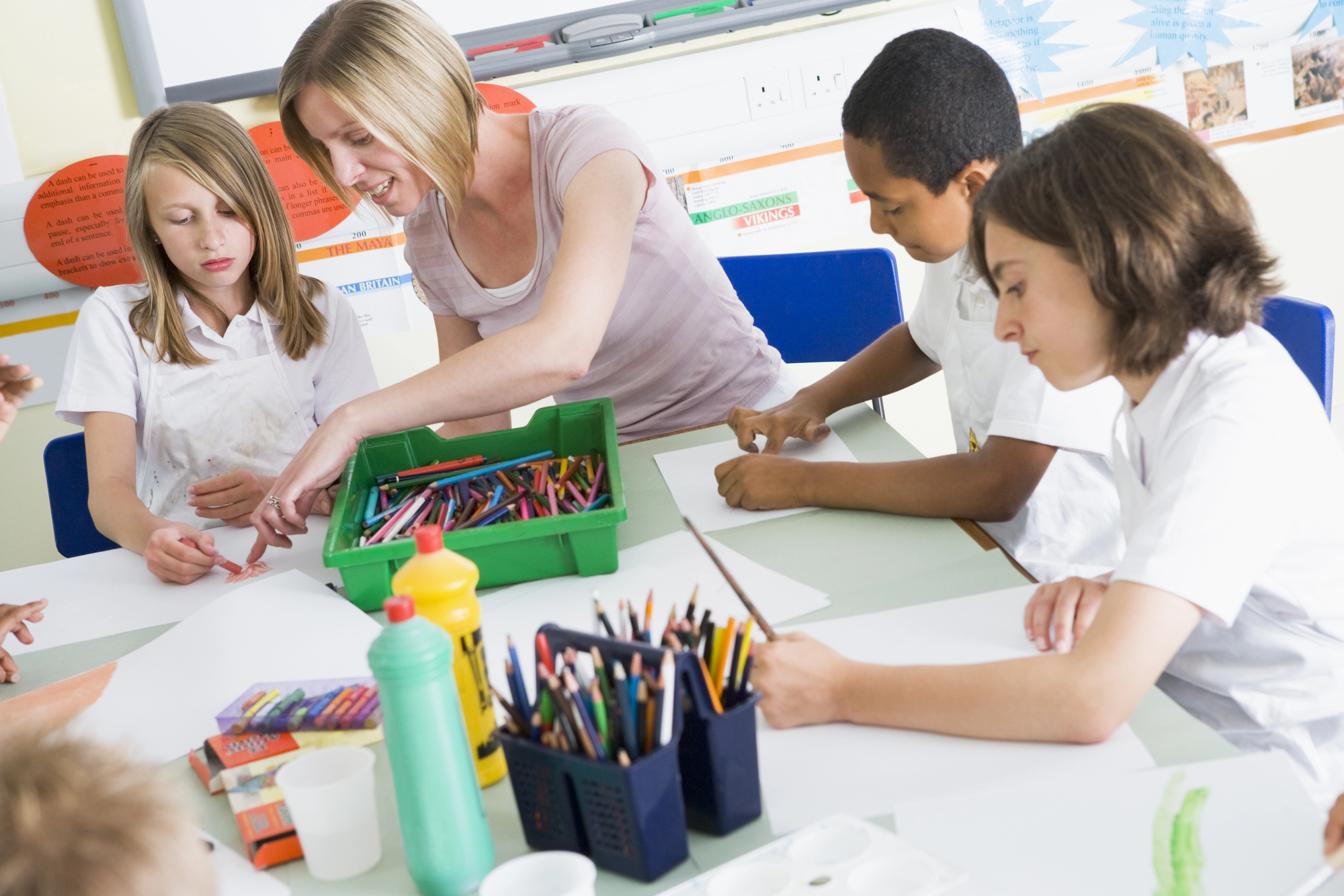 A teacher assists a group of children seated around a table in a classroom. The table is filled with art supplies, including crayons, colored pencils, paint, and paper. The children are engaged in a creative activity.