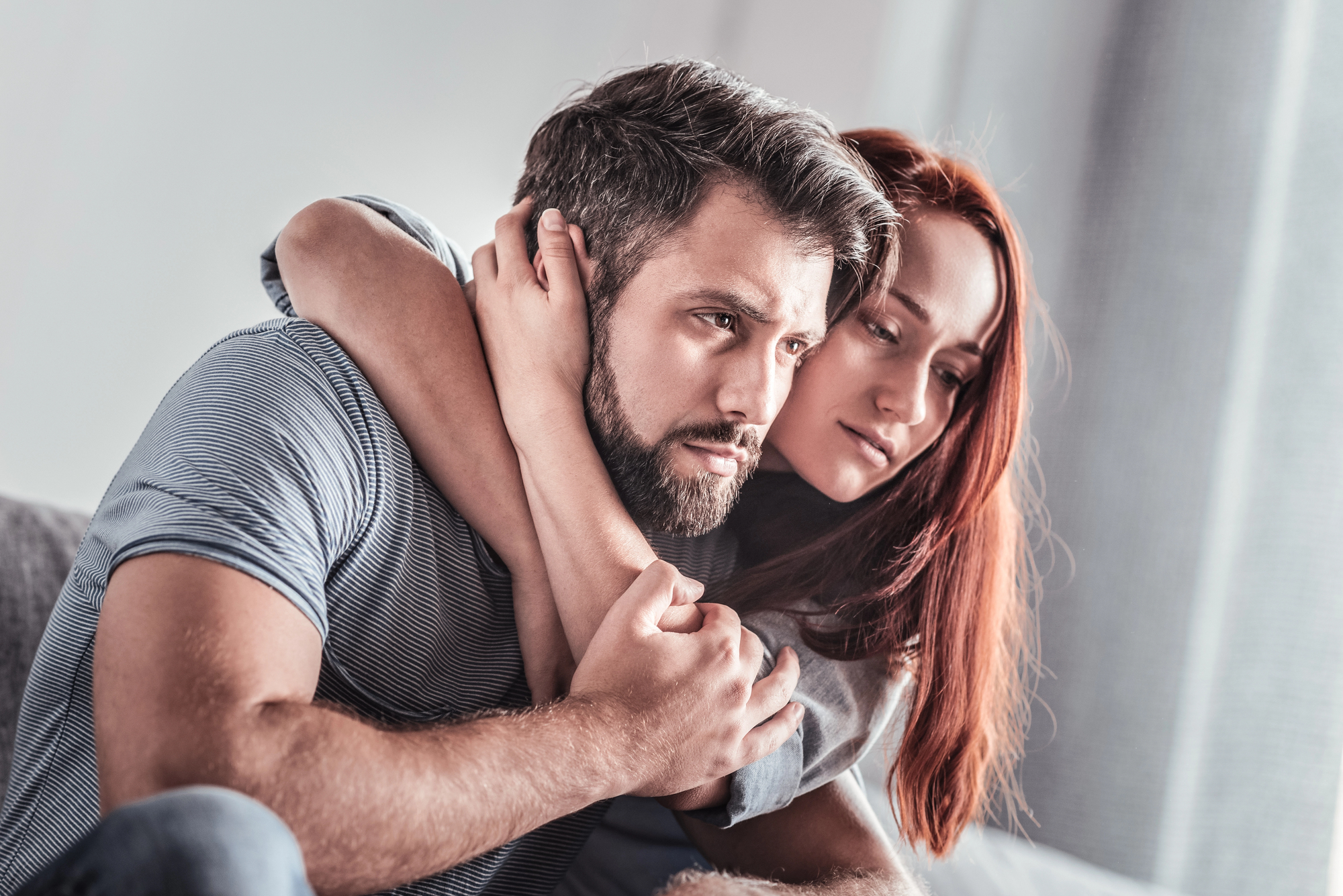 A couple sits closely on a sofa, with the woman embracing the man from behind. They both have serious expressions, gazing thoughtfully in the same direction. The background is softly blurred, suggesting an indoor setting.