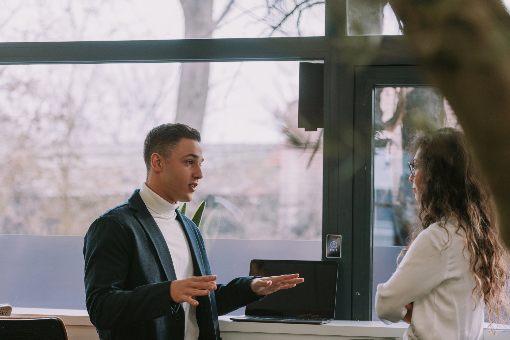 A man and a woman are having a conversation in an office setting. The man gestures while speaking, and the woman listens attentively. A laptop is open on the table in front of them. Large windows show a blurred outdoor view.