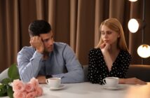 A man and woman sit at a table with cups of coffee in front of them. Both appear contemplative or concerned. The table has a bouquet of pink roses. The background features brown curtains and soft, round lights.