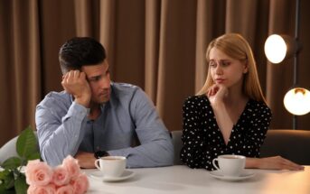 A man and woman sit at a table with cups of coffee in front of them. Both appear contemplative or concerned. The table has a bouquet of pink roses. The background features brown curtains and soft, round lights.