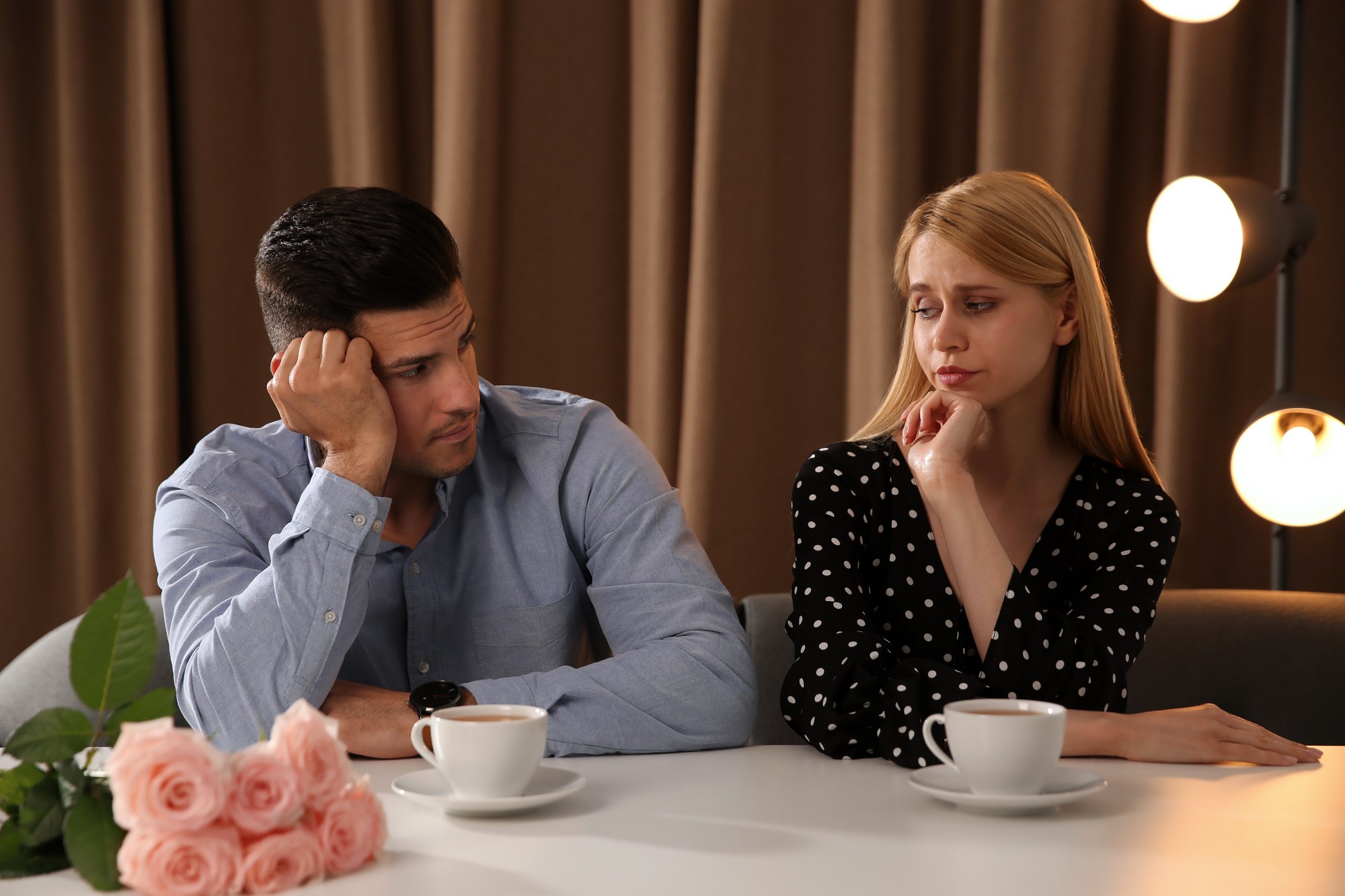 A man and woman sit at a table with cups of coffee in front of them. Both appear contemplative or concerned. The table has a bouquet of pink roses. The background features brown curtains and soft, round lights.