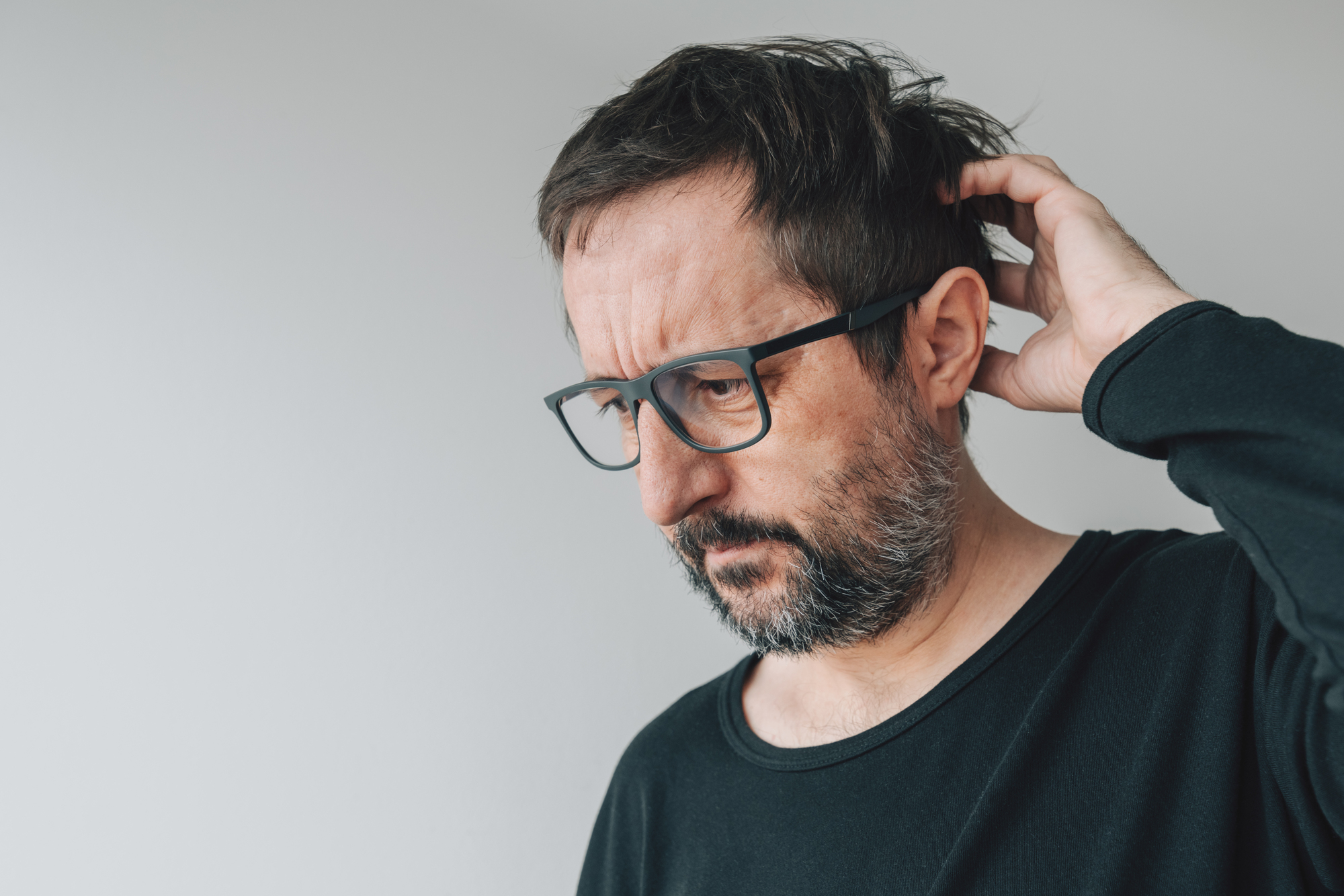 A man with glasses and a beard, wearing a black shirt, is scratching his head while looking downward. He appears to be deep in thought or pondering something, standing against a plain white background.