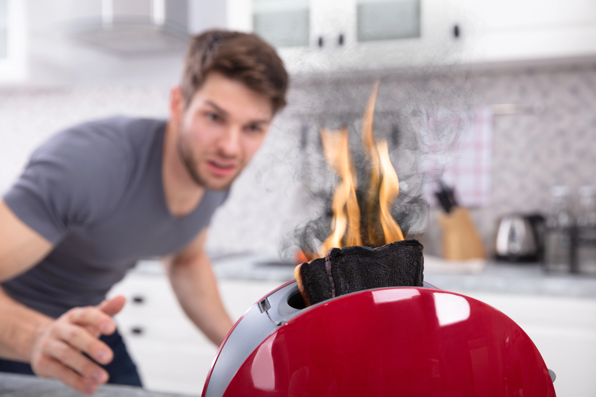 A man in a kitchen stares in shock at a red toaster with flames and smoke coming from burnt toast. The background shows kitchen counters and cabinets.