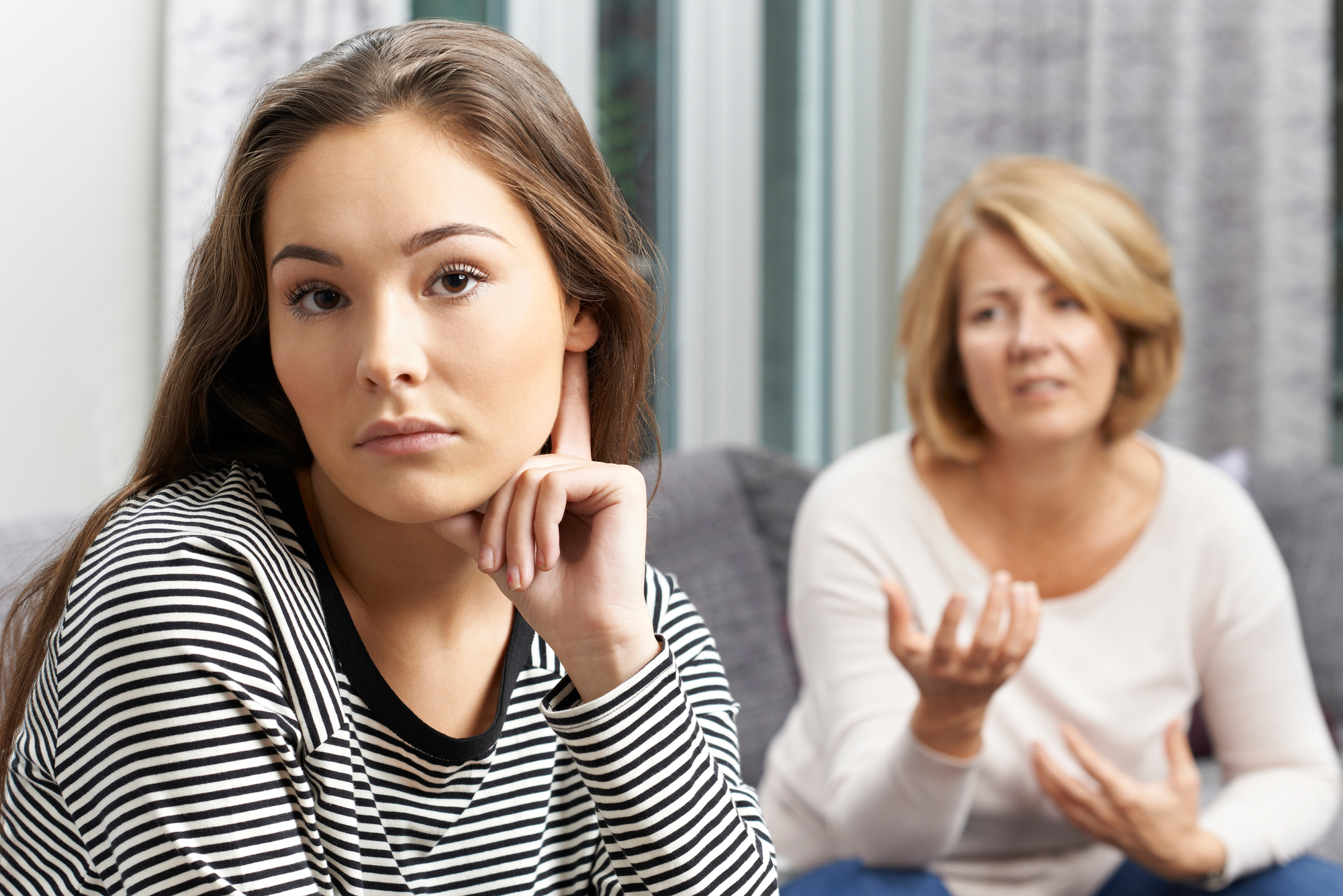 A young woman with long hair wearing a striped shirt looks pensively at the camera, resting her chin on her hand. In the background, an older woman with short hair gestures as if speaking, seated on a couch in a softly lit room.