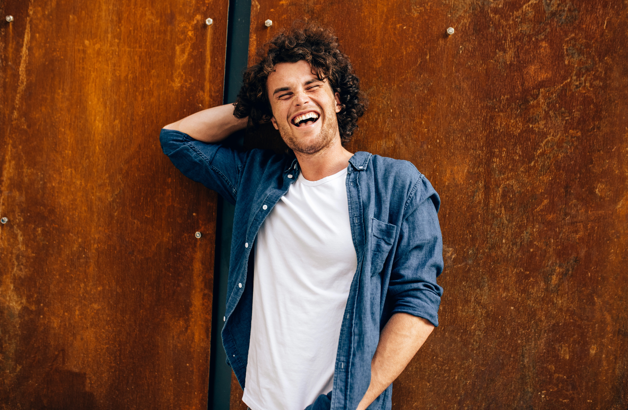 A man with curly hair wearing a white t-shirt and an open denim shirt stands in front of a rust-colored wall, laughing with one hand behind his head.