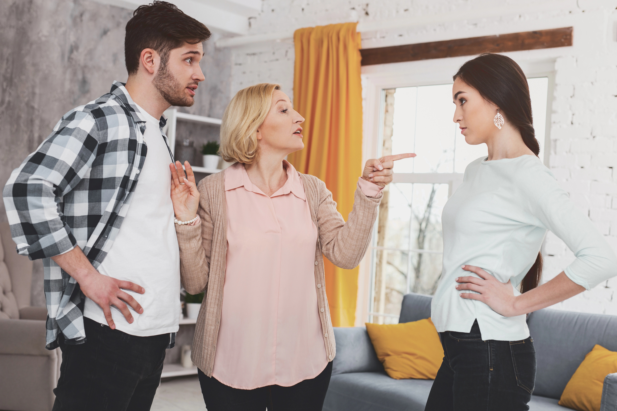 A woman gestures and speaks to a young man and woman standing in a living room. The young man, wearing a plaid shirt, looks surprised. The young woman, in a white sweater, has her hands on her hips. There's a couch and yellow curtains in the background.