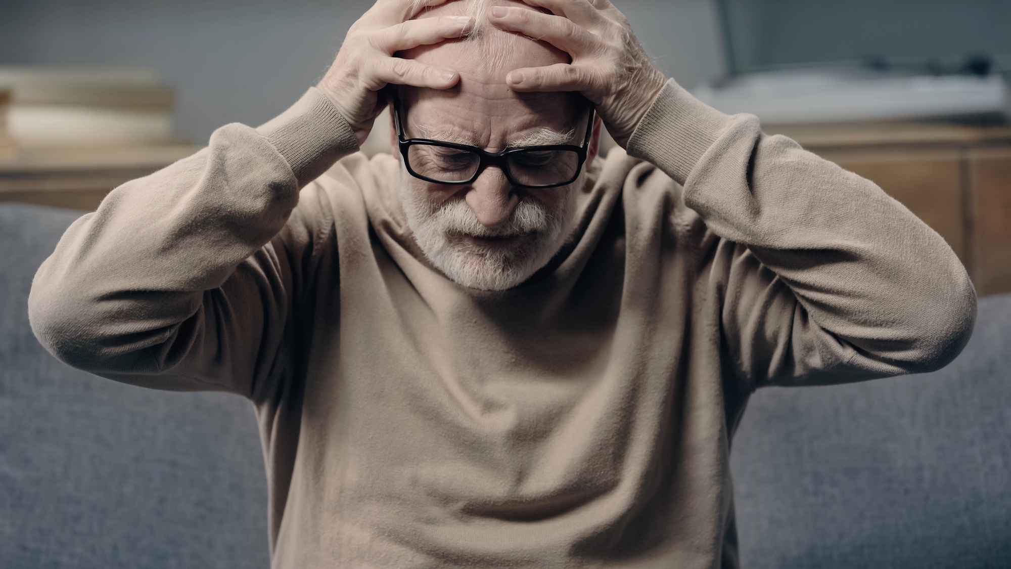 An elderly man with glasses sits on a couch, holding his head with both hands, appearing distressed or deep in thought. He wears a beige sweater and is in a softly lit room.