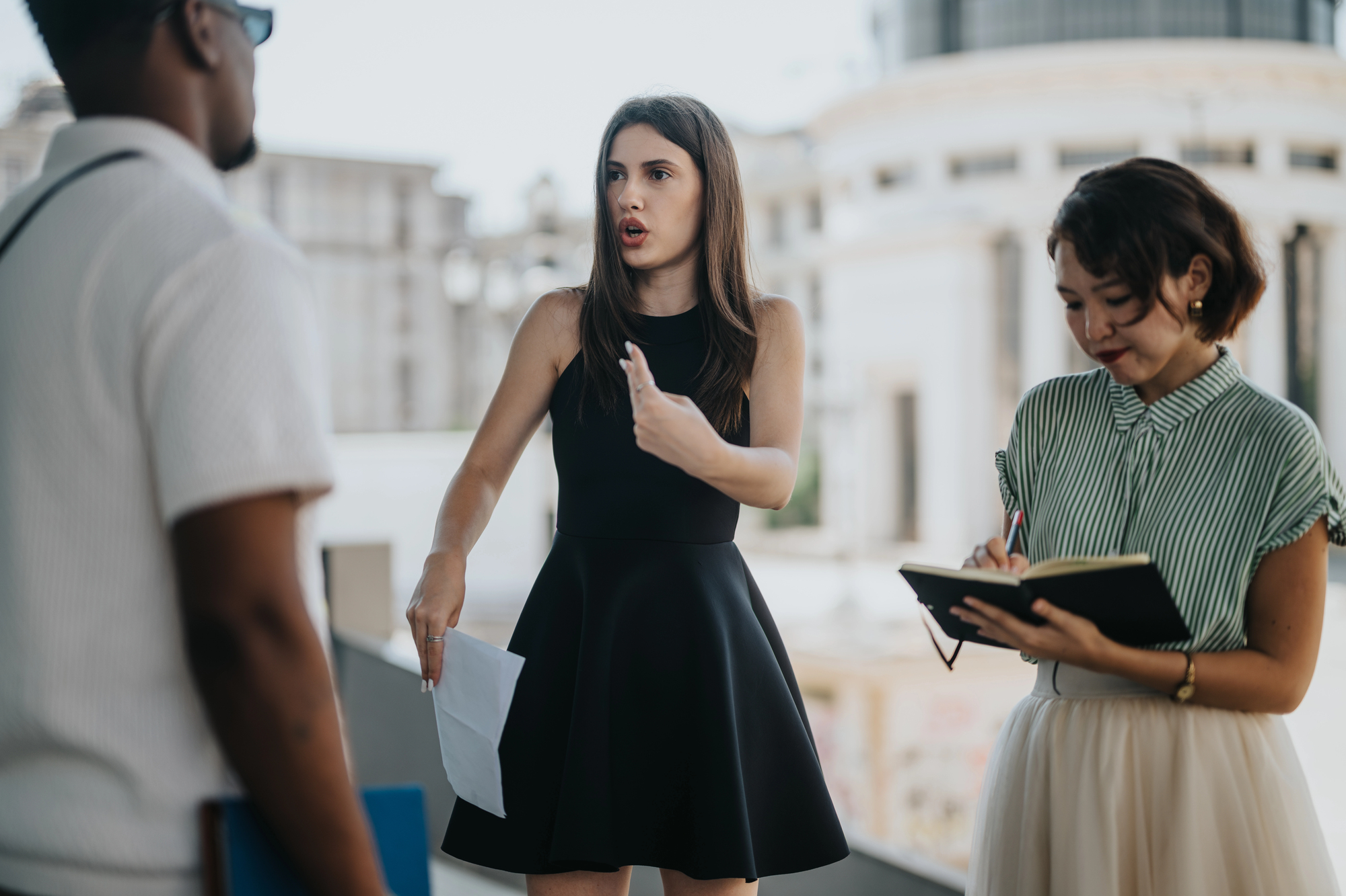 A woman in a black dress is animatedly speaking to a man, while another woman in a striped shirt takes notes. They are outdoors on an urban balcony with blurred buildings in the background.