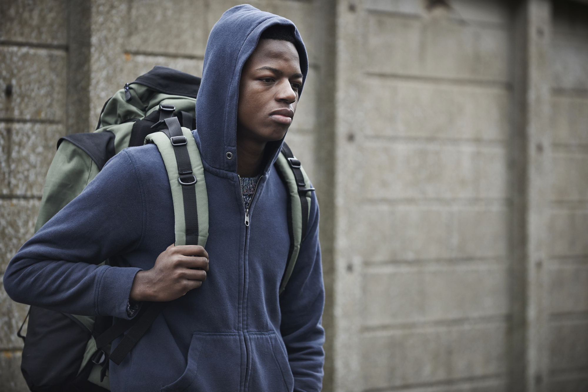 A person wearing a blue hoodie and carrying a large backpack walks past a concrete wall. They appear deep in thought and the scene suggests a journey or travel.