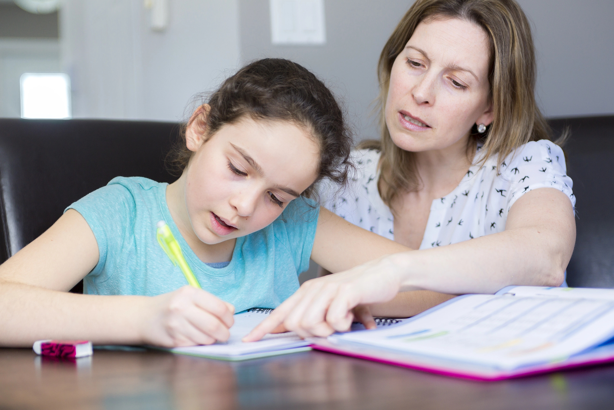 A woman and a young girl are sitting at a table. The girl is writing with a pen in a notebook, while the woman, pointing at the notebook, looks on attentively. They appear to be engaged in a learning or homework activity.