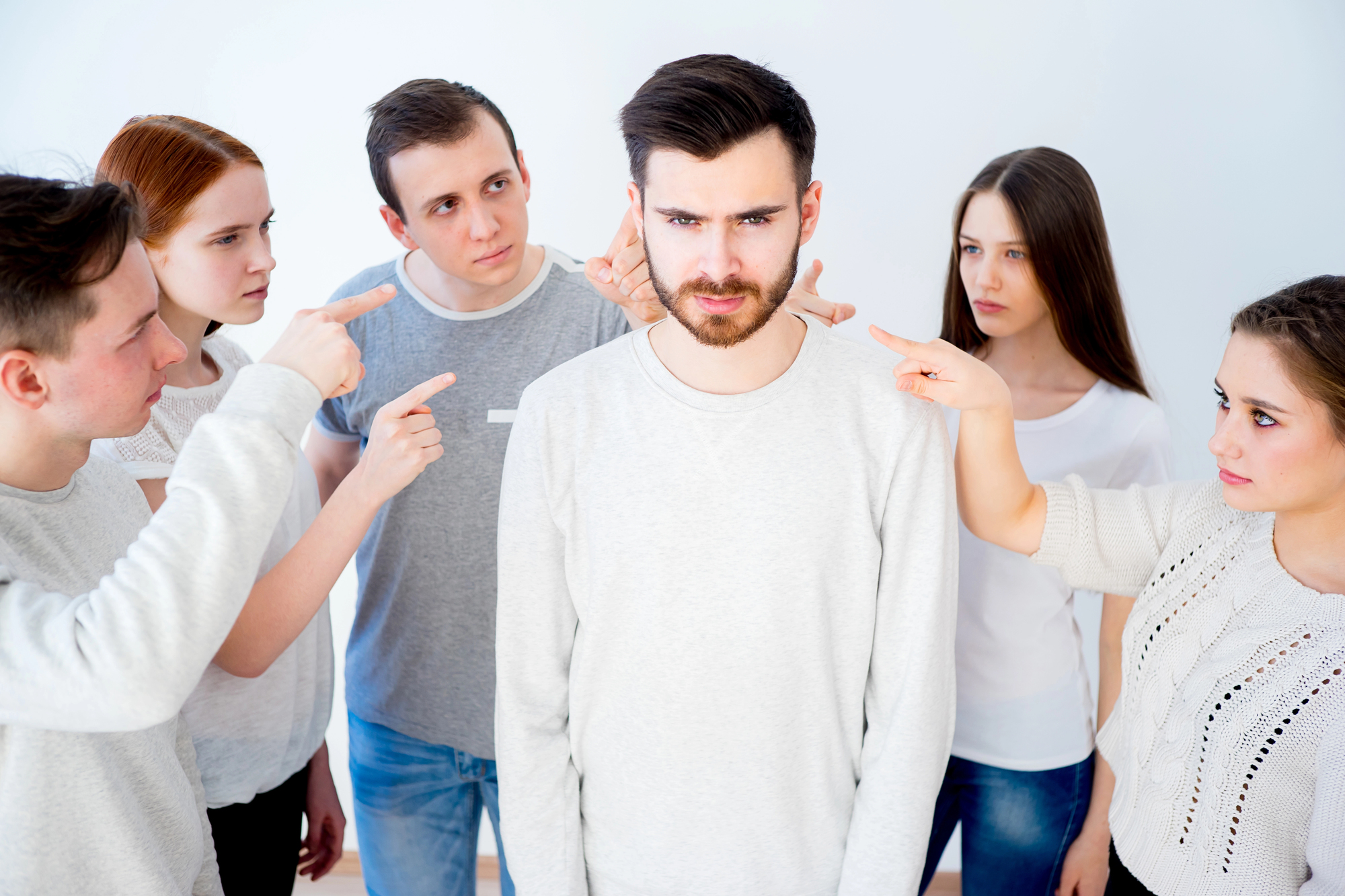 A man stands in the center looking serious, surrounded by five people pointing fingers at him. The background is a plain white wall. The group appears to be in an intense discussion or confrontation.