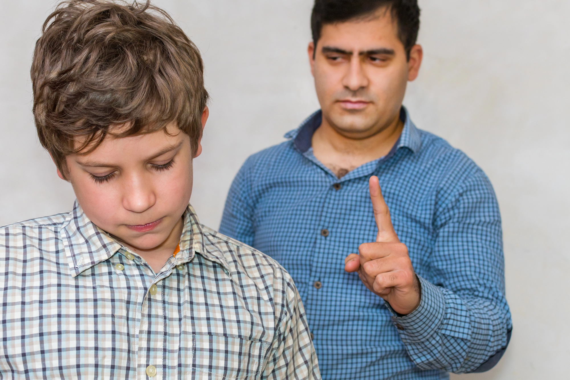 A man in a blue plaid shirt gestures with his index finger raised, while a boy in a checkered shirt looks down with a serious expression. The background is plain and neutral.