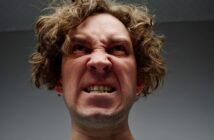 A person with curly hair and a scrunched-up face, showing their teeth, appears to be expressing anger or frustration. They are looking upward, and the background is a plain, grayish color.