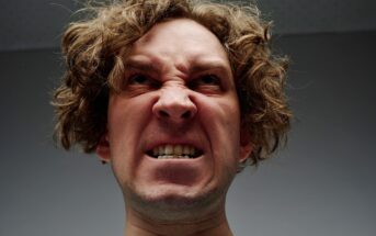 A person with curly hair and a scrunched-up face, showing their teeth, appears to be expressing anger or frustration. They are looking upward, and the background is a plain, grayish color.