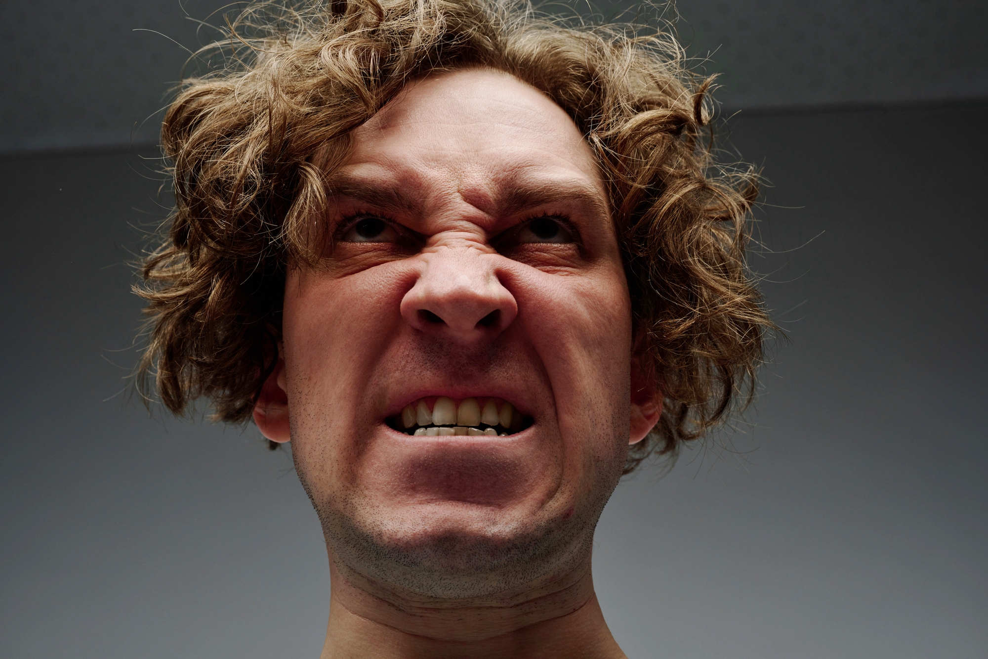 A person with curly hair and a scrunched-up face, showing their teeth, appears to be expressing anger or frustration. They are looking upward, and the background is a plain, grayish color.