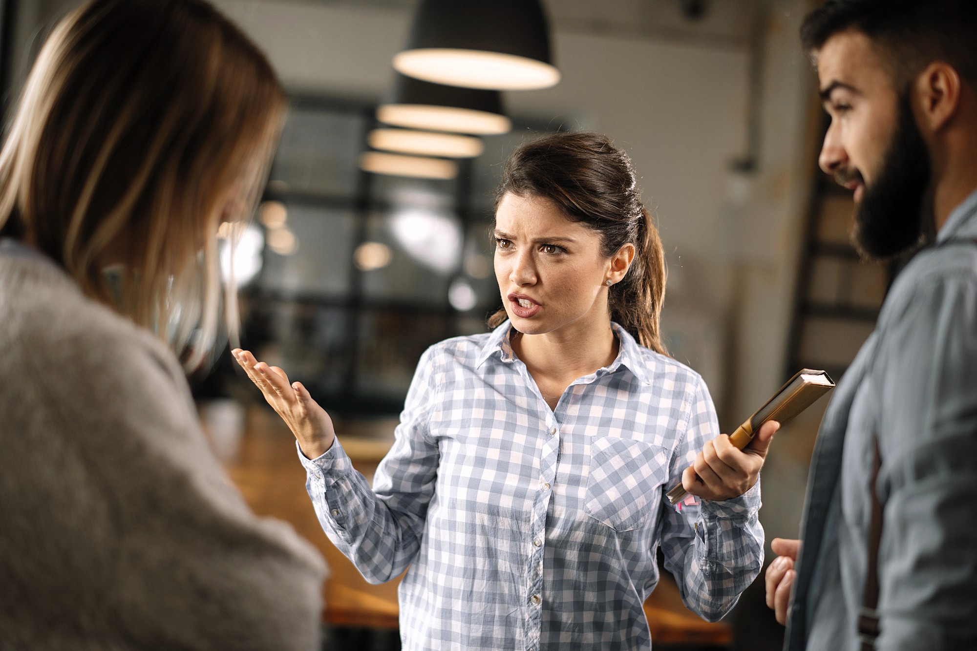 A woman in a checkered shirt is gesturing and speaking emphatically to two colleagues in an office setting. One person faces away while the other listens attentively. The background shows blurred industrial lighting and decor.