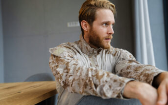 A man with a beard and light brown hair sits at a wooden table, wearing a camouflage-patterned jacket. He gazes into the distance with a thoughtful expression. The background shows gray walls and a window with light curtains.