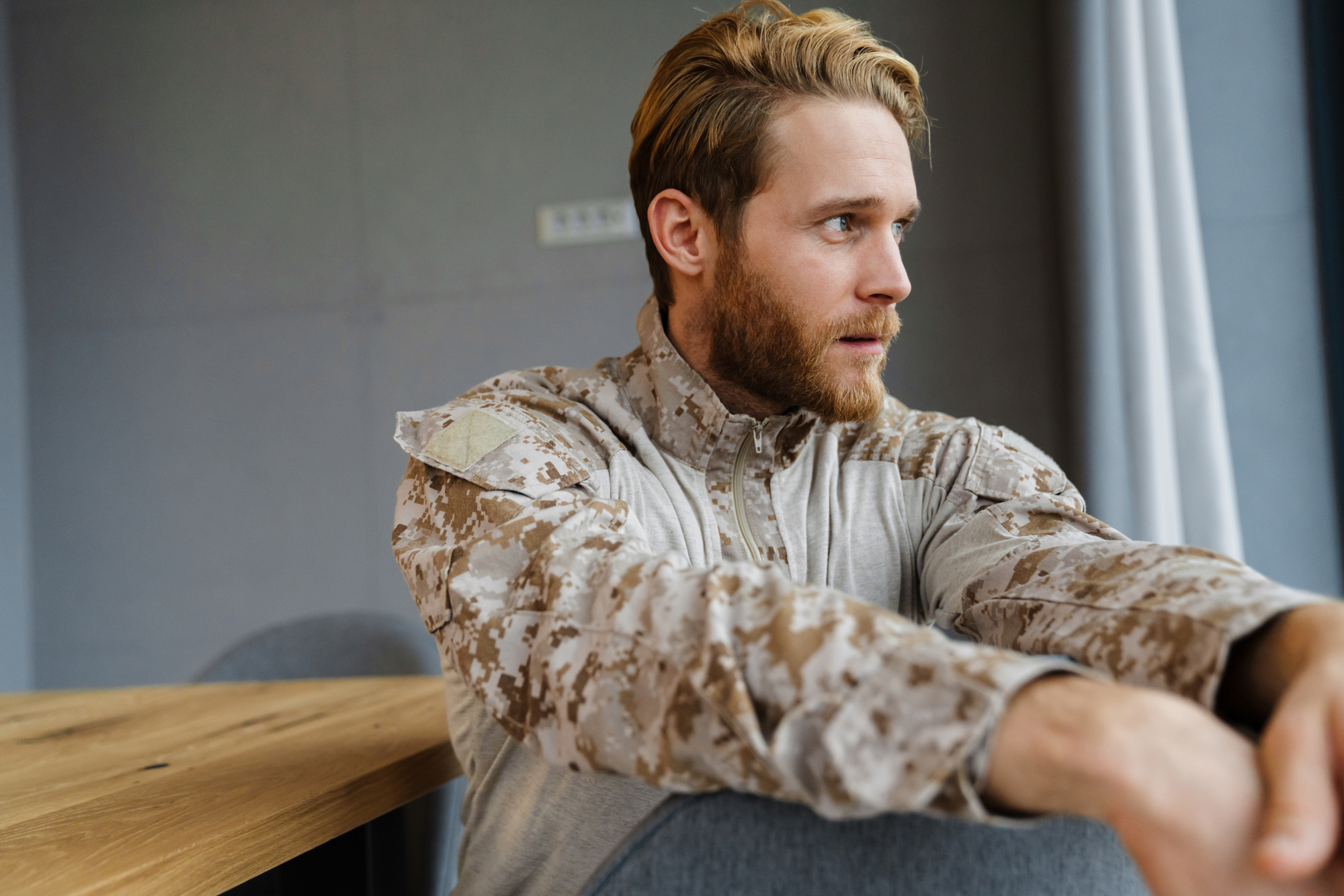 A man with a beard and light brown hair sits at a wooden table, wearing a camouflage-patterned jacket. He gazes into the distance with a thoughtful expression. The background shows gray walls and a window with light curtains.