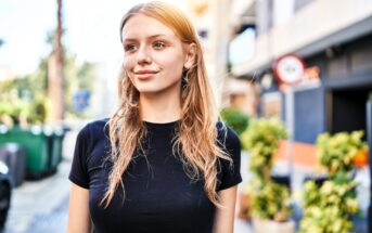 A person with long hair wearing a black shirt stands outdoors on a sunny day. They are smiling slightly, and the background shows a street with buildings and greenery.