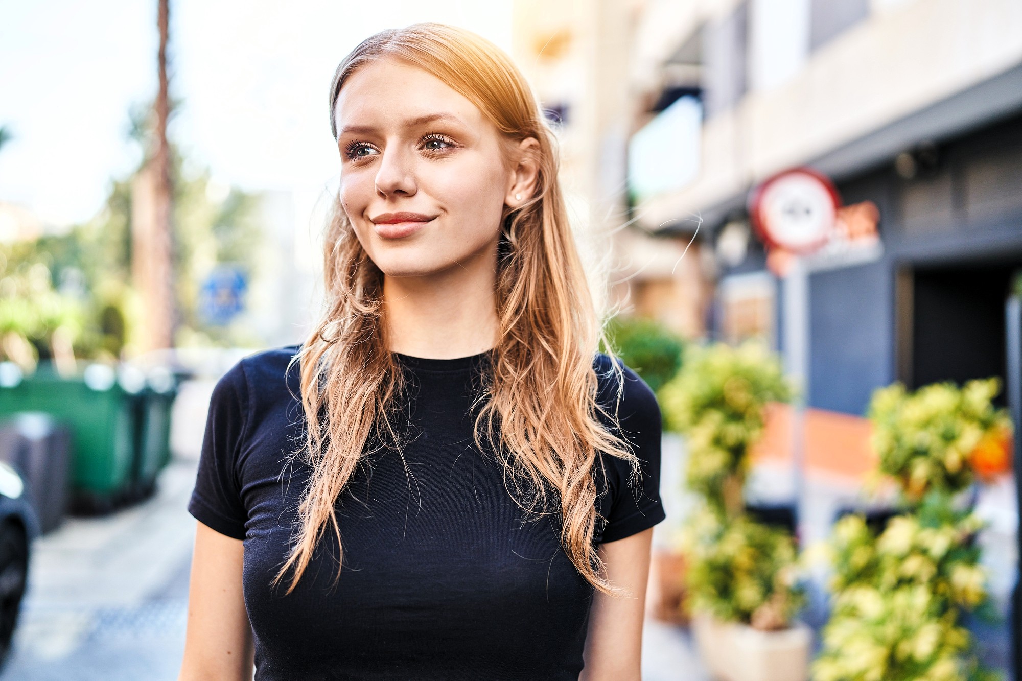 A person with long hair wearing a black shirt stands outdoors on a sunny day. They are smiling slightly, and the background shows a street with buildings and greenery.