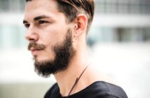 A person with a beard, mustache, and short hair is in profile, wearing a black shirt. They have multiple earrings and a nose ring. The background is blurred with an indistinct urban setting.