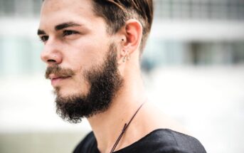 A person with a beard, mustache, and short hair is in profile, wearing a black shirt. They have multiple earrings and a nose ring. The background is blurred with an indistinct urban setting.