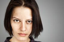 Close-up of a person with short, dark hair and a serious expression, looking directly at the camera. The background is a neutral gray.