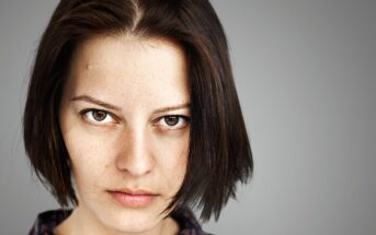 Close-up of a person with short, dark hair and a serious expression, looking directly at the camera. The background is a neutral gray.