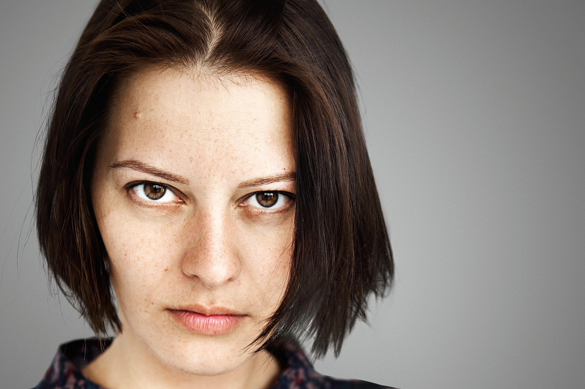 Close-up of a person with short, dark hair and a serious expression, looking directly at the camera. The background is a neutral gray.