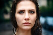 A woman with wet hair and a serious expression looks directly at the camera. She is outdoors, with a blurred background of greenery and a building. The focus is on her face, emphasizing her eyes and features.