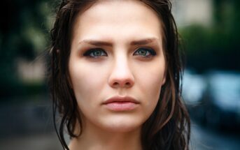 A woman with wet hair and a serious expression looks directly at the camera. She is outdoors, with a blurred background of greenery and a building. The focus is on her face, emphasizing her eyes and features.