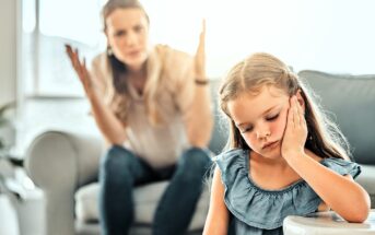 A young girl with long hair looks upset, resting her chin in her hand, sitting on a couch. In the background, a woman appears to be talking or explaining something with her hands raised, seated on another couch. Both are at home.