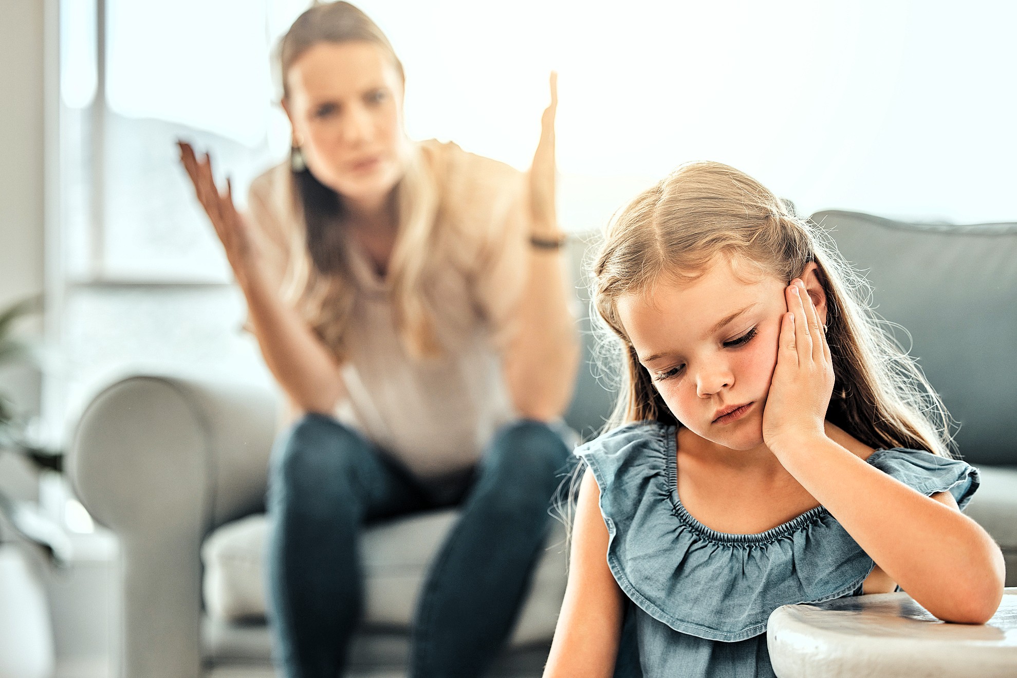 A young girl with long hair looks upset, resting her chin in her hand, sitting on a couch. In the background, a woman appears to be talking or explaining something with her hands raised, seated on another couch. Both are at home.