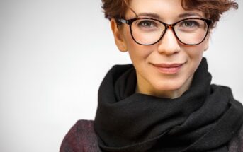 A person with short, curly hair and glasses, wearing a black scarf and a dark top, smiles slightly. The background is a plain, light color.