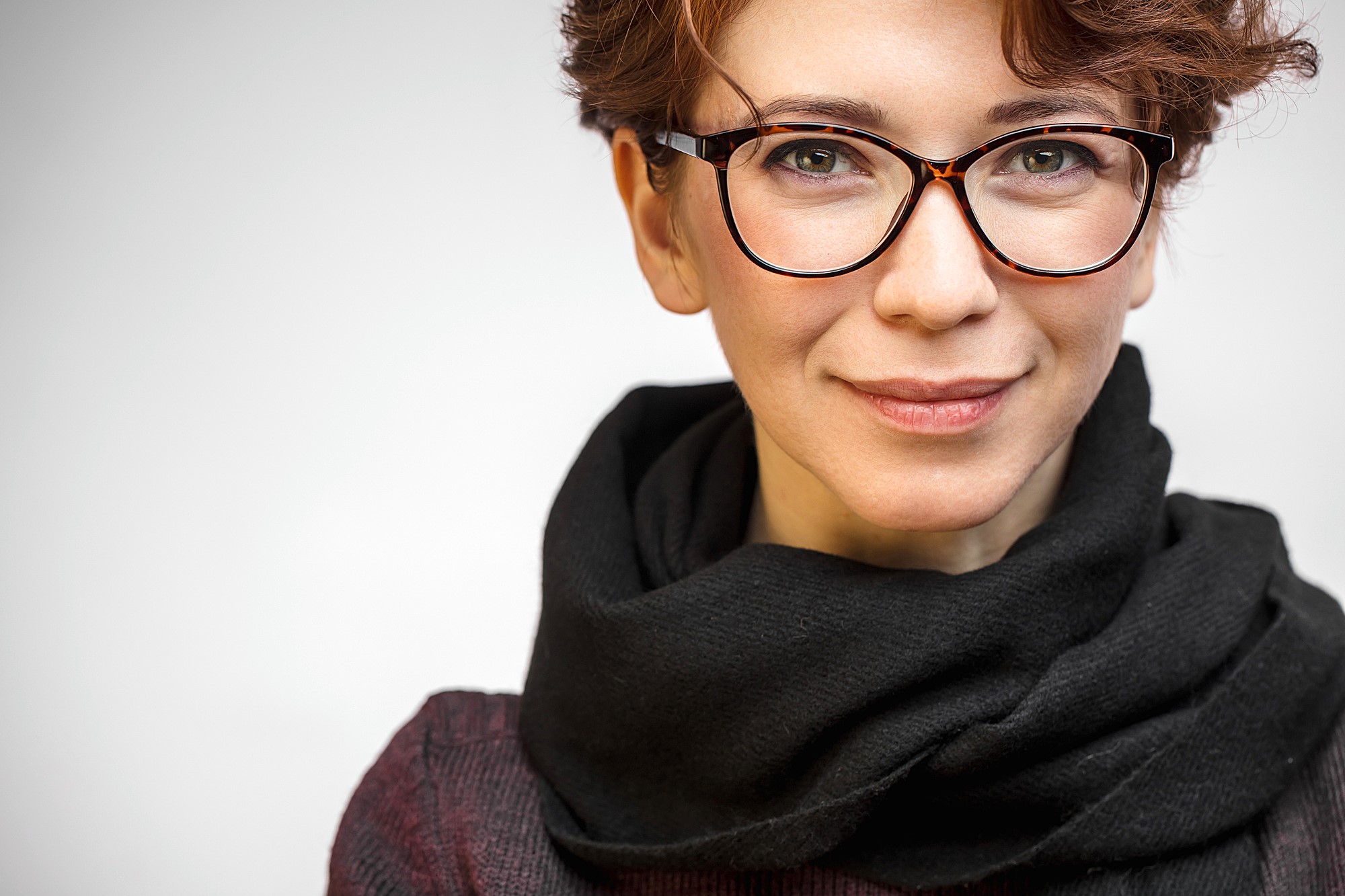 A person with short, curly hair and glasses, wearing a black scarf and a dark top, smiles slightly. The background is a plain, light color.