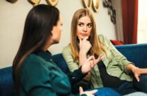 Two women sitting on a blue couch have a serious conversation. The woman on the right, with long blonde hair, looks thoughtful with her hand on her chin, while the woman on the left gestures expressively.