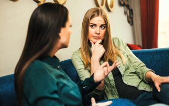 Two women sitting on a blue couch have a serious conversation. The woman on the right, with long blonde hair, looks thoughtful with her hand on her chin, while the woman on the left gestures expressively.