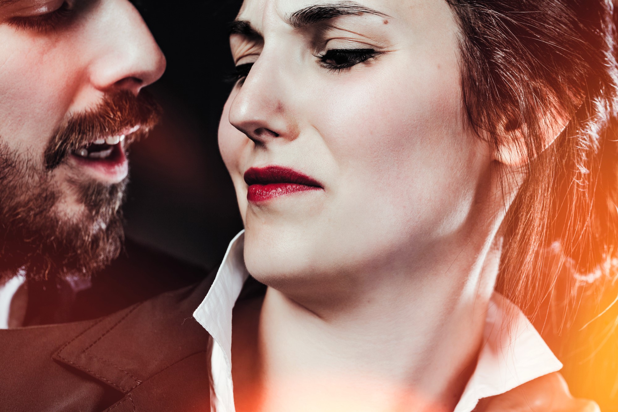 A close-up of a woman in a suit with a skeptical expression, slightly turning away from a man with a beard who is speaking to her. The lighting creates a warm, dramatic effect around them.