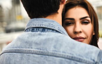 A woman with long brown hair is being embraced by a person wearing a denim jacket, seen from behind. Her face is partially visible over their shoulder, and she has a serene expression. The background is softly blurred.