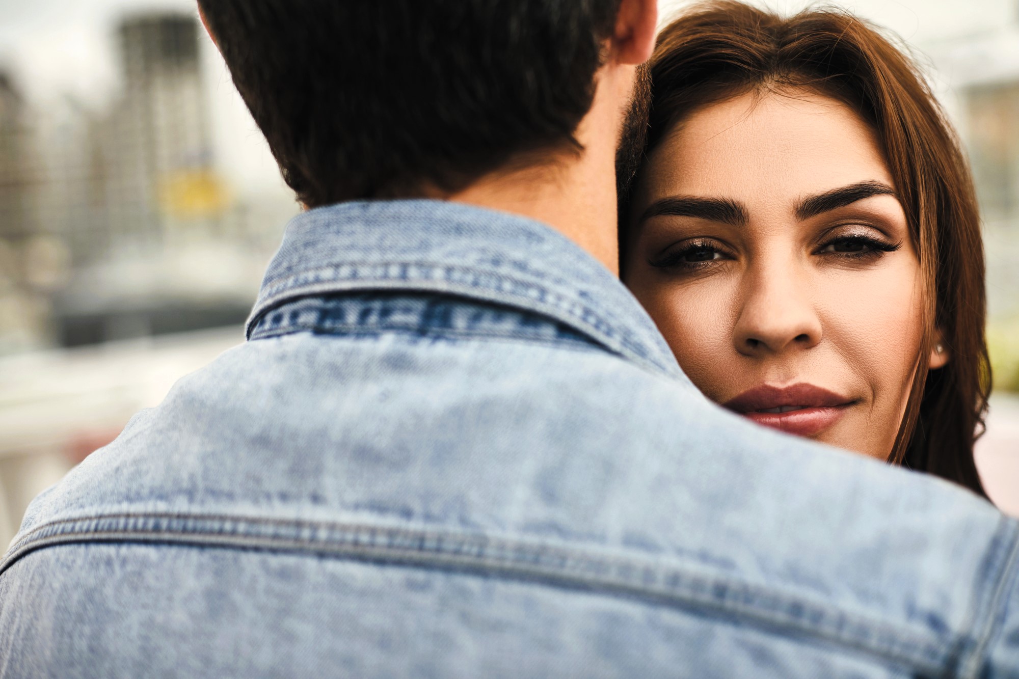 A woman with long brown hair is being embraced by a person wearing a denim jacket, seen from behind. Her face is partially visible over their shoulder, and she has a serene expression. The background is softly blurred.