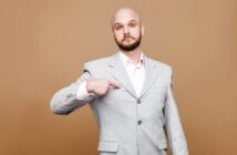 Bald man with a beard, wearing a light gray suit over a white shirt, points at himself with a serious expression, standing against a plain brown background.