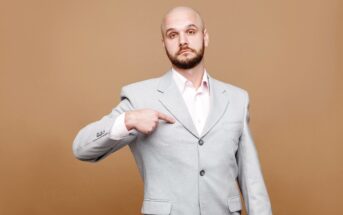 Bald man with a beard, wearing a light gray suit over a white shirt, points at himself with a serious expression, standing against a plain brown background.