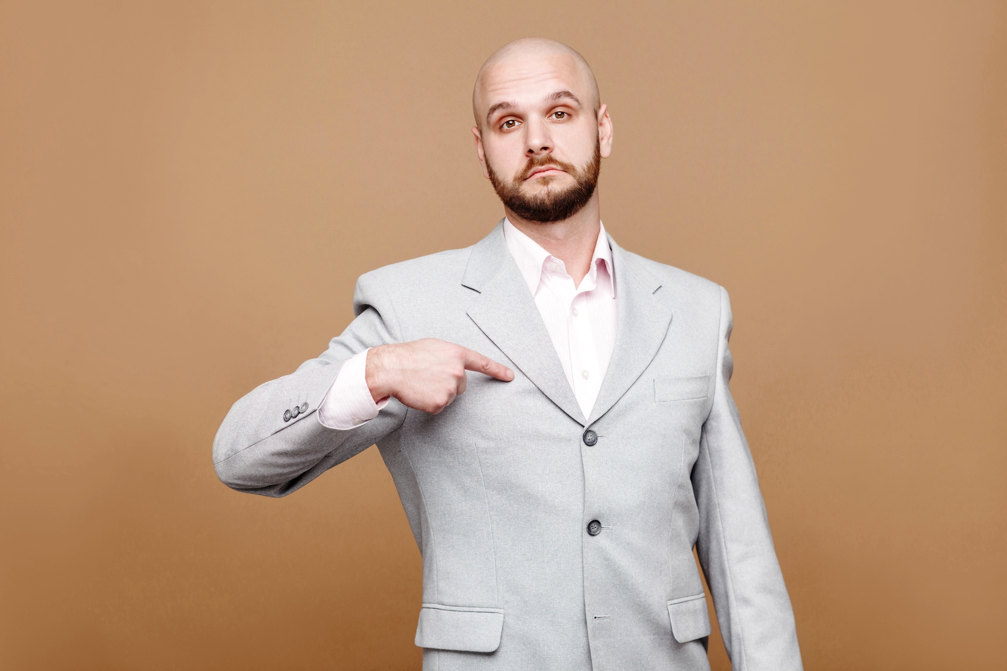 Bald man with a beard, wearing a light gray suit over a white shirt, points at himself with a serious expression, standing against a plain brown background.