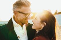 A man and woman smiling at each other in sunlight. The man, with glasses and short hair, wears a dark coat and white scarf. The woman, with long hair, wears a red jacket. The sun is setting behind them, creating a warm glow.