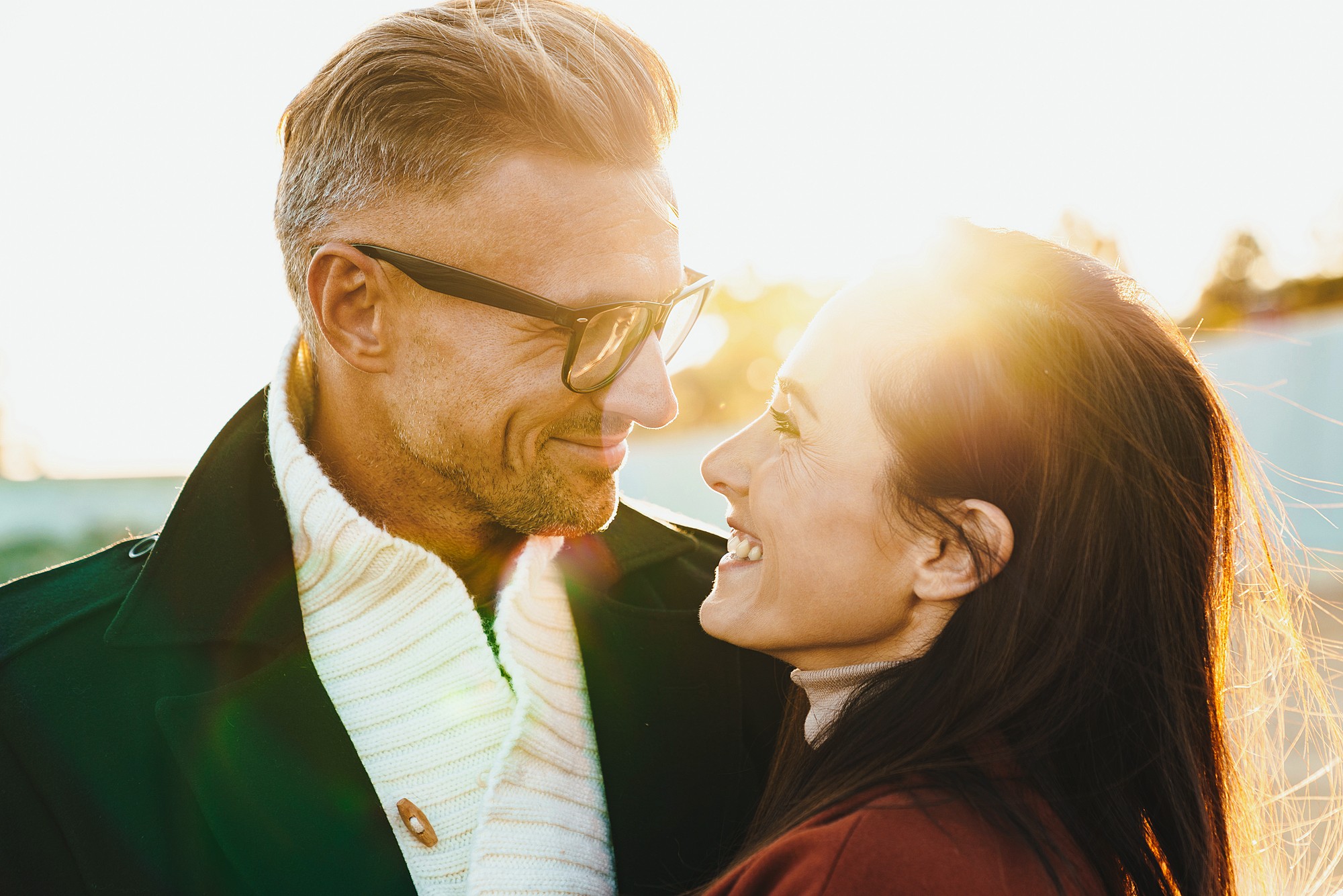 A man and woman smiling at each other in sunlight. The man, with glasses and short hair, wears a dark coat and white scarf. The woman, with long hair, wears a red jacket. The sun is setting behind them, creating a warm glow.